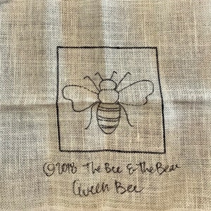 K&ouml;nnte beinhalten: Ein gesticktes Design in Schwarzwei&szlig; einer Biene auf einem beigefarbenen Stoff mit dem Text "From the bee to the bear with bee".