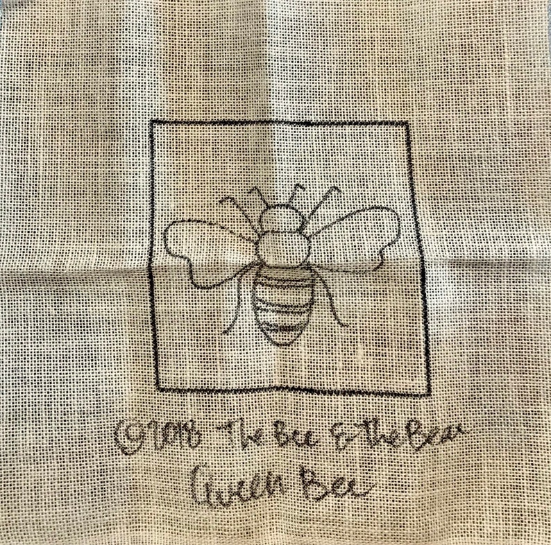 K&ouml;nnte beinhalten: Ein gesticktes Design in Schwarzwei&szlig; einer Biene auf einem beigefarbenen Stoff mit dem Text "From the bee to the bear with bee".