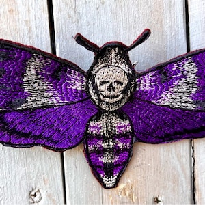 May include: Purple and black embroidered moth patch with a skull design. The patch is iron-on and measures approximately 3 inches wide.