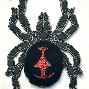 May include: Black spider embroidered patch with a red hourglass on its abdomen. The patch is made of felt and has a black outline.