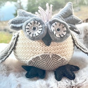 May include: A gray and white knitted owl with large black eyes and a brown and white patterned body. The owl is sitting on a white fluffy surface.