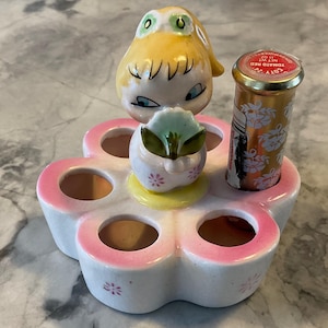 Könnte beinhalten: Ein weißer, blumenförmiger Lippenstiftständer aus Keramik mit sechs Fächern. Eine kleine Mädchenfigur mit gelben Haaren und einem grünen Kleid hält ein grünes Blatt und sitzt auf dem Ständer. Ein goldener Lippenstifttubus mit roter Kappe und weißem Blumendruck befindet sich in einem der Fächer. Der Tubus trägt die Aufschrift "COTY - TOMATO RED - NET WT. 1/10 OZ."