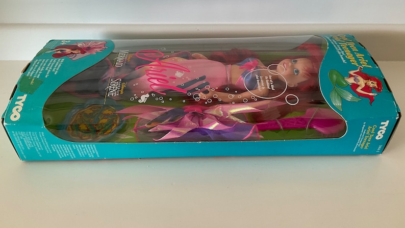 Pode incluir: Uma boneca Ariel rosa e azul do filme da Disney A Pequena Sereia. A boneca est&aacute; usando uma cauda de sereia rosa e azul e tem cabelo comprido e vermelho. A caixa apresenta o texto "Cool Tween Ariel Teenager" e "Disney's The Little Mermaid Sirene Supreme".
