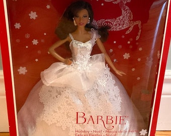 May include: A Barbie doll wearing a white gown with lace detailing and a silver bodice. The doll is standing in a red box with snowflakes and a reindeer design. The box also has the text "25th Anniversary" and "Barbie" on it.