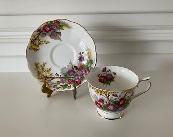 May include: A white teacup and saucer with a floral pattern featuring purple and pink flowers, yellow branches, and gold accents.