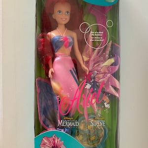 Pode incluir: Uma boneca sereia rosa e azul com cabelo ruivo e um top azul e rosa. A boneca est&aacute; segurando uma pizza de algas marinhas rosa e azul. A caixa diz "Cool Teen Ariel Ariel Teenager" e "Disney's The Little Mermaid La Petite Sirene".