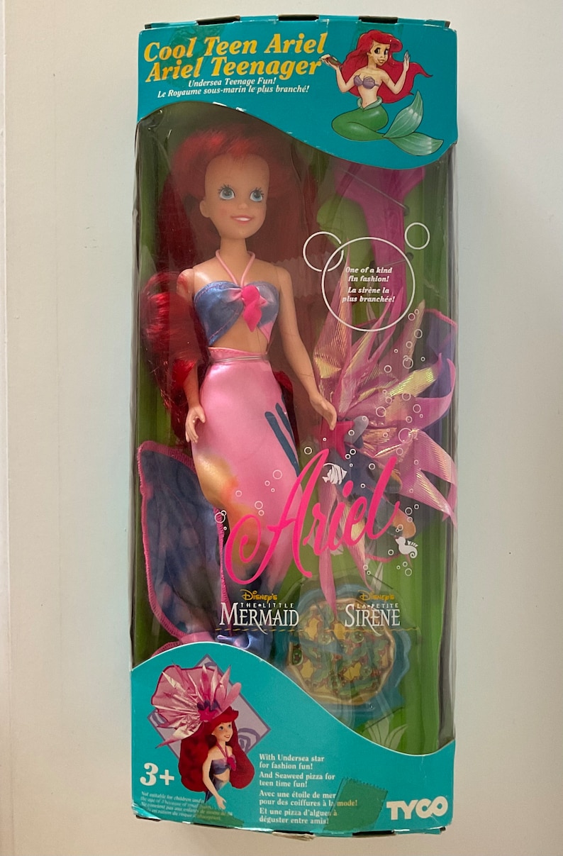 Pode incluir: Uma boneca sereia rosa e azul com cabelo ruivo e um top azul e rosa. A boneca est&aacute; segurando uma pizza de algas marinhas rosa e azul. A caixa diz "Cool Teen Ariel Ariel Teenager" e "Disney's The Little Mermaid La Petite Sirene".
