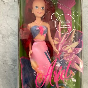 Pode incluir: Uma boneca de pl&aacute;stico da Ariel do filme da Disney A Pequena Sereia. Ela est&aacute; usando uma cauda de sereia rosa e azul e um top azul e rosa. A boneca est&aacute; embalada em uma caixa de pl&aacute;stico transparente com um fundo rosa e azul. A caixa tamb&eacute;m inclui uma concha rosa e uma cauda de sereia azul e rosa.