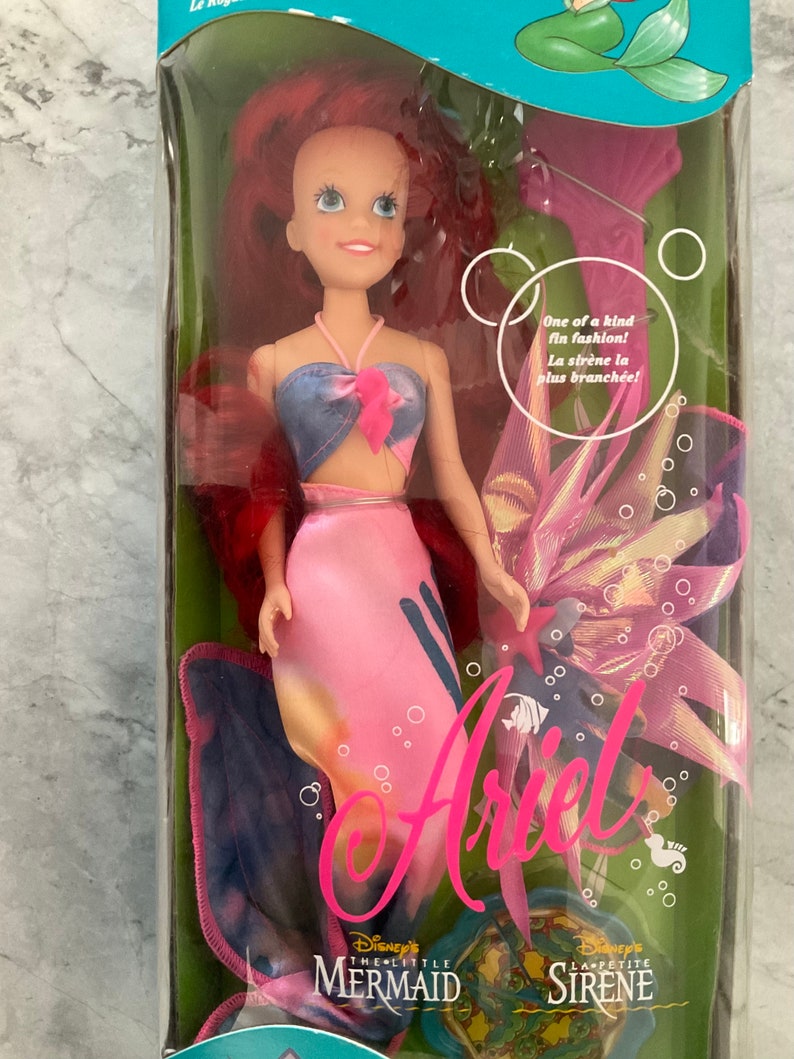 Pode incluir: Uma boneca de pl&aacute;stico da Ariel do filme da Disney A Pequena Sereia. Ela est&aacute; usando uma cauda de sereia rosa e azul e um top azul e rosa. A boneca est&aacute; embalada em uma caixa de pl&aacute;stico transparente com um fundo rosa e azul. A caixa tamb&eacute;m inclui uma concha rosa e uma cauda de sereia azul e rosa.