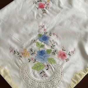 May include: White tablecloth with a yellow border and embroidered floral designs in shades of blue, pink, yellow, and green. The tablecloth features a delicate lace trim.
