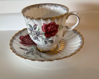 May include: A white teacup and saucer set with a red rose design. The cup and saucer have gold trim and a scalloped edge. The teacup has a handle and sits on a small pedestal base. The saucer has a matching rose design.