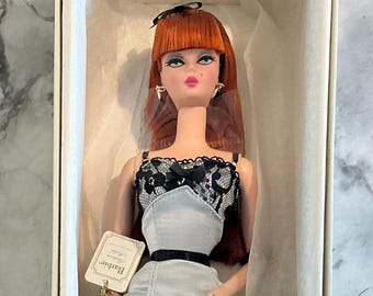 May include: A Barbie doll in a box. The doll has red hair, blue eyes, and is wearing a gray and black lace outfit. The doll is in a white box with a tag that says "Barbie".
