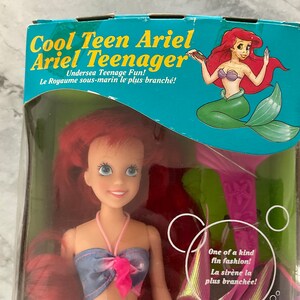 Pode incluir: Uma boneca Ariel de cabelo ruivo com um top de biqu&iacute;ni azul e rosa e uma barbatana de cauda rosa. A boneca est&aacute; embalada em uma caixa com uma imagem de desenho animado de Ariel na frente. A caixa diz "Cool Teen Ariel Ariel Teenager Undersea Teenage Fun! Le Royaume sous-marin le plus branch&eacute;!" e "One of a kind fin fashion! La sir&egrave;ne la plus branch&eacute;e!"