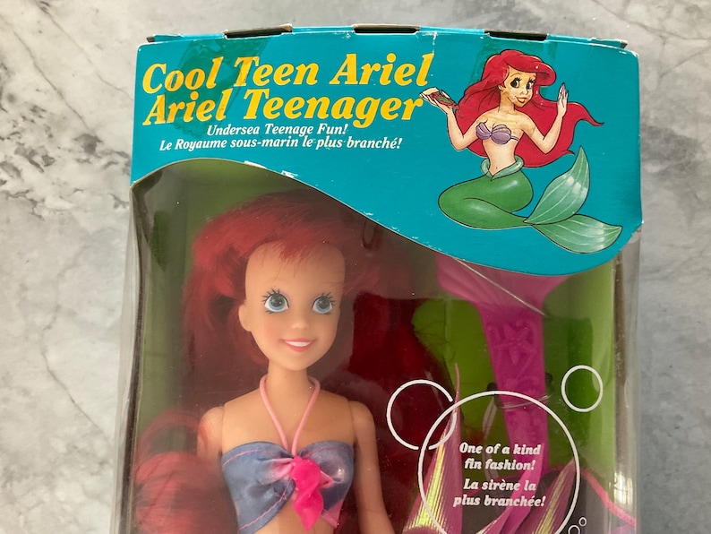 Pode incluir: Uma boneca Ariel de cabelo ruivo com um top de biqu&iacute;ni azul e rosa e uma barbatana de cauda rosa. A boneca est&aacute; embalada em uma caixa com uma imagem de desenho animado de Ariel na frente. A caixa diz "Cool Teen Ariel Ariel Teenager Undersea Teenage Fun! Le Royaume sous-marin le plus branch&eacute;!" e "One of a kind fin fashion! La sir&egrave;ne la plus branch&eacute;e!"