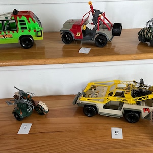 Peut inclure: Cinq véhicules jouets, dont une Jeep Jurassic Park verte, une Jeep rouge et grise avec un canon, une Jeep camouflage avec une cage, une moto verte et une Jeep jaune avec un filet. Les véhicules sont numérotés de 1 à 5.