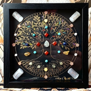 Puede incluir: Una obra de arte enmarcada con un árbol de la vida con detalles en pan de oro y varias piedras preciosas de colores. El árbol está rodeado por un círculo de piedras más pequeñas sobre un fondo negro.