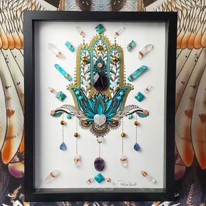 May include: A framed artwork featuring a turquoise and gold Hamsa hand with crystals and beads. The artwork is signed "Paint & Bead".