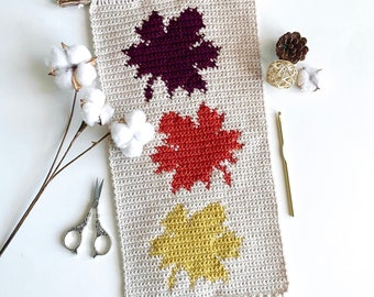 May include: A crocheted wall hanging with three fall leaves in shades of burgundy, orange, and yellow. The leaves are crocheted in a simple, geometric style. The wall hanging has a fringe at the bottom and is attached to a piece of driftwood.