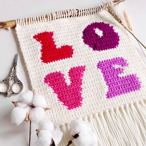 Puede incluir: Un tapiz de pared de crochet blanco con la palabra "LOVE" escrita en hilo rosa, rojo y morado. El tapiz tiene un borde de flecos y se muestra con tallos de algodón.