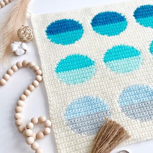 May include: A white crocheted wall hanging with a geometric pattern of blue and gray circles. The hanging has a brown tassel attached to the bottom.