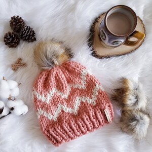 Puede incluir: Un gorro de punto rosa y blanco con un patrón de chevron y un pompón esponjoso en la parte superior. El gorro tiene una pequeña etiqueta de cuero marrón con el texto "Warm & Cozy" en ella.