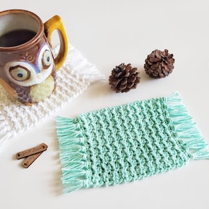 May include: A set of two crocheted coasters with fringe. One coaster is white and the other is light green. The coasters are sitting on a white surface. The text "Love & Stitch" is on two wooden tags.