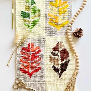 May include: Crocheted wall hanging featuring four leaf designs in fall colors: green, yellow, red, and brown. The wall hanging is cream-colored with gray squares and a fringe. A wooden dowel and wooden beads are attached.