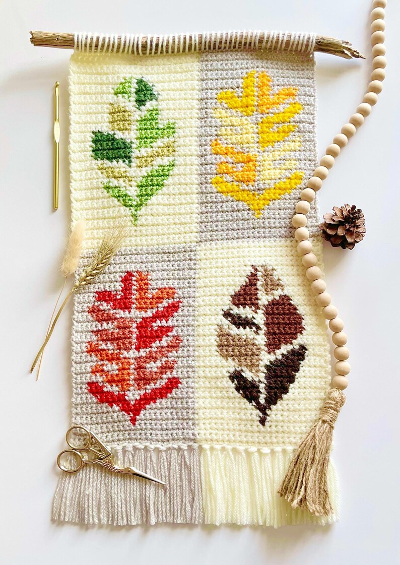 May include: Crocheted wall hanging featuring four leaf designs in fall colors: green, yellow, red, and brown. The wall hanging is cream-colored with gray squares and a fringe. A wooden dowel and wooden beads are attached.