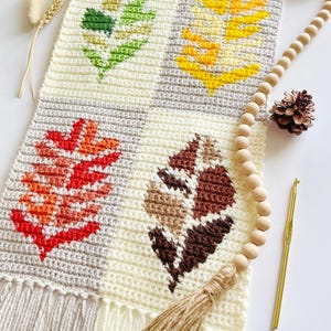May include: A handmade crochet wall hanging featuring four leaf designs in green, yellow, red, and brown. The leaves are set against a cream and gray background, with a fringed bottom and a wooden dowel for hanging. A wooden bead garland and a crochet hook are also present.