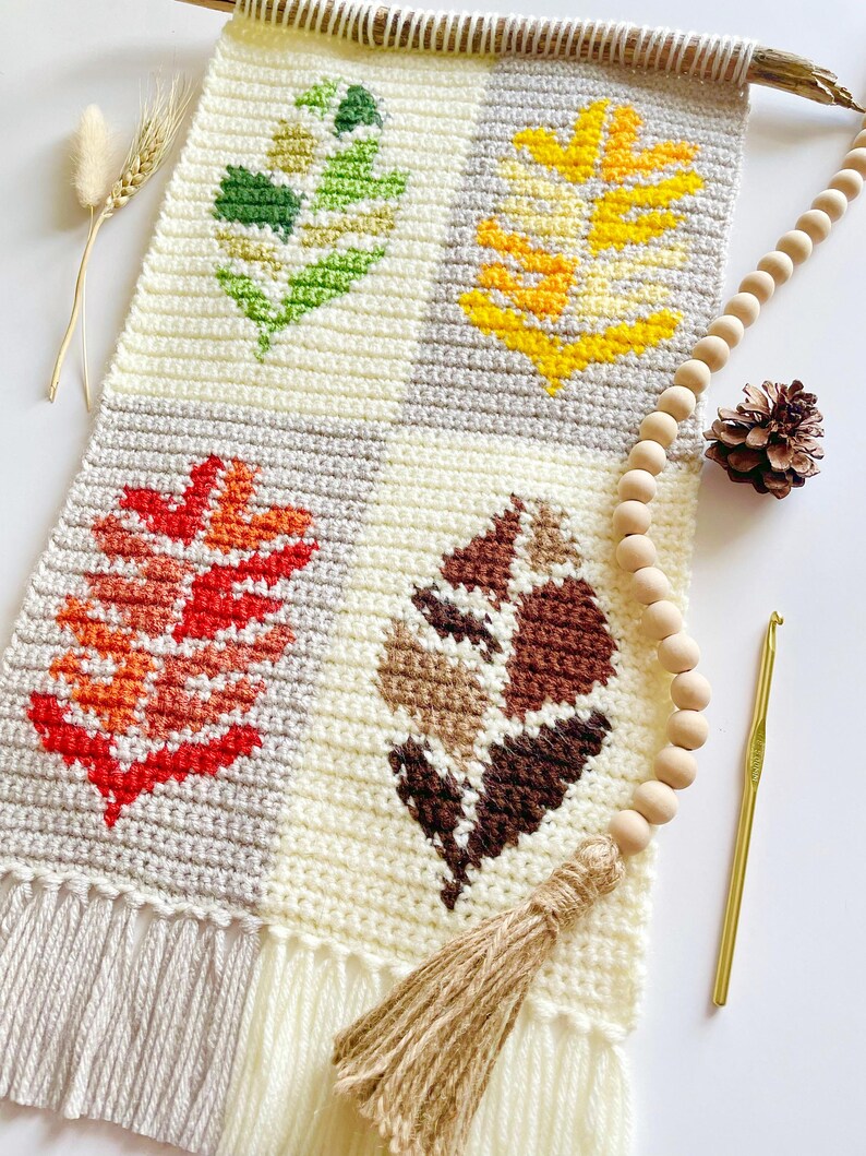May include: A handmade crochet wall hanging featuring four leaf designs in green, yellow, red, and brown. The leaves are set against a cream and gray background, with a fringed bottom and a wooden dowel for hanging. A wooden bead garland and a crochet hook are also present.