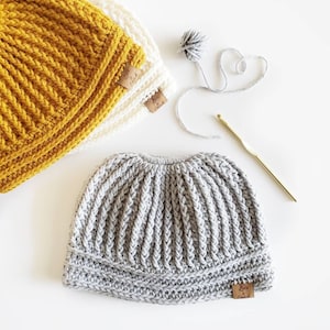 May include: Three crocheted hats in different colors: mustard yellow, white, and gray. The hats have a ribbed texture and a brown leather label with the text "Love & Sew" on them.