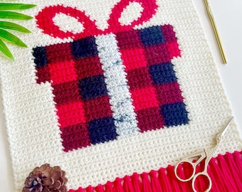 May include: A crocheted wall hanging featuring a red and black plaid gift box with a white bow. The hanging has a red fringe border and is displayed on a white surface.