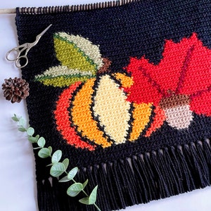 May include: Black tapestry with a fall harvest design. The tapestry features a pumpkin, maple leaf, and acorn in shades of orange, red, green, and beige. The tapestry has a fringe at the bottom and is hung from a wooden dowel.