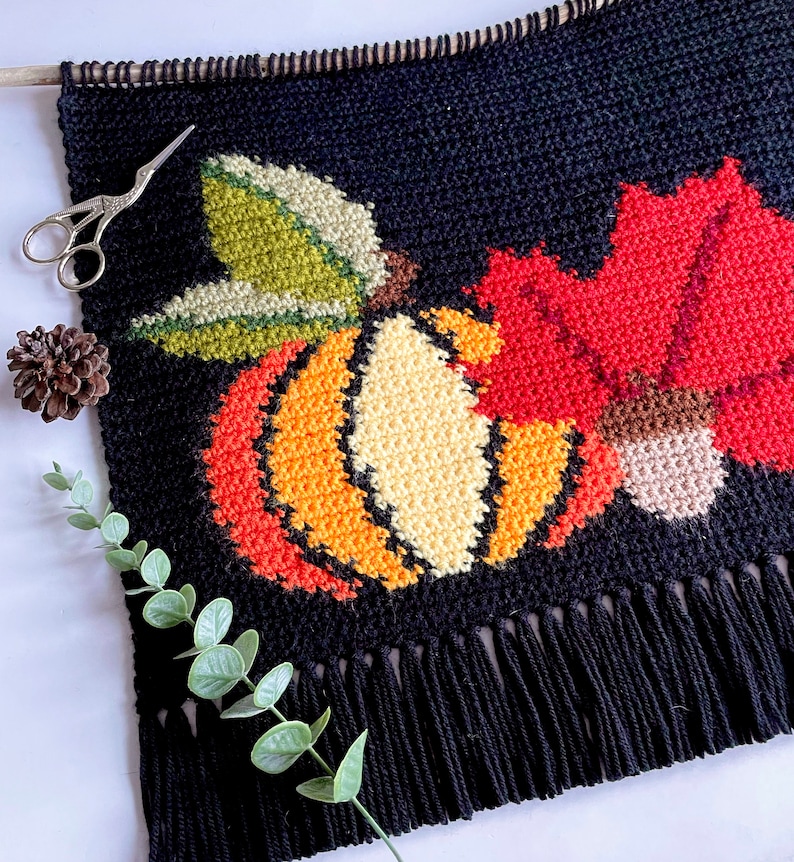 May include: Black tapestry with a fall harvest design. The tapestry features a pumpkin, maple leaf, and acorn in shades of orange, red, green, and beige. The tapestry has a fringe at the bottom and is hung from a wooden dowel.
