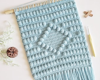 May include: A light blue crocheted wall hanging with a textured pattern and fringe. The hanging is attached to a wooden dowel rod.