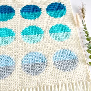 May include: A white crocheted wall hanging with a fringe border features a pattern of nine ombre circles in shades of blue and gray.