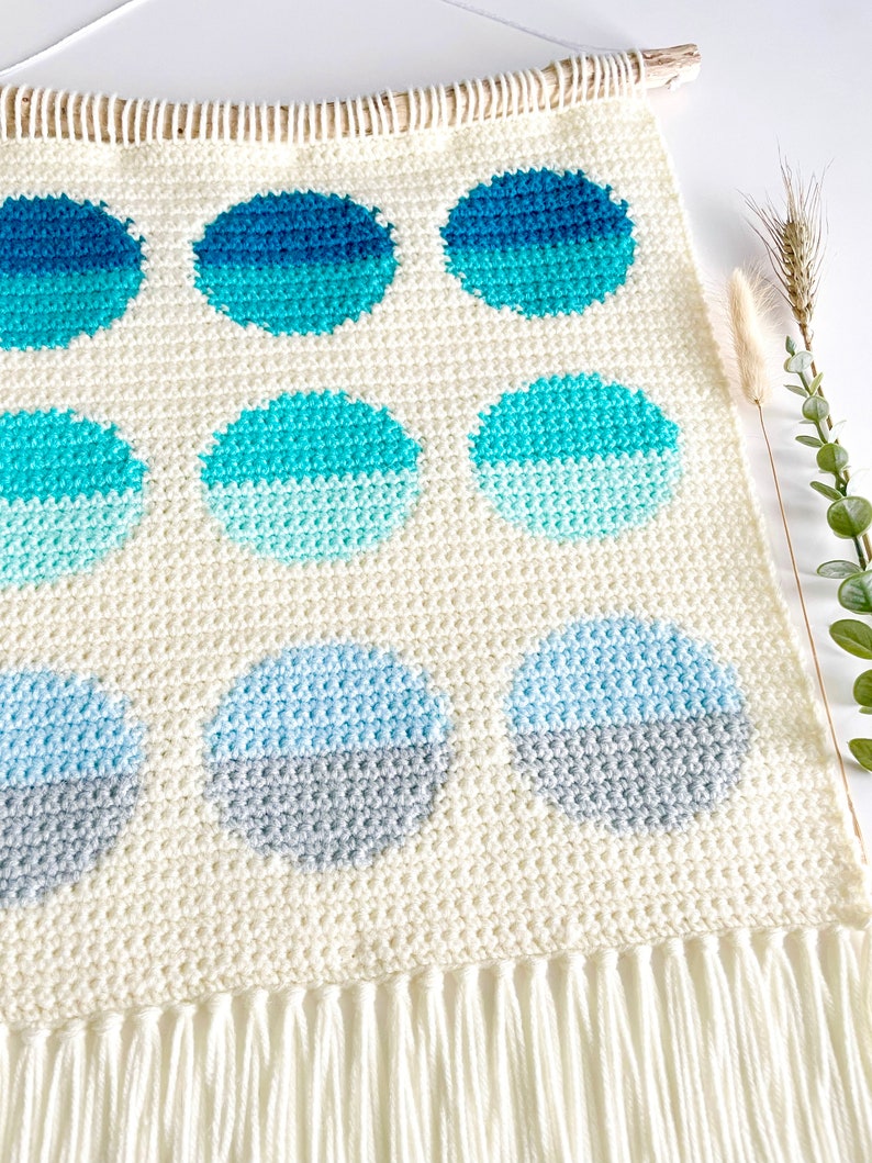 May include: A white crocheted wall hanging with a fringe border features a pattern of nine ombre circles in shades of blue and gray.
