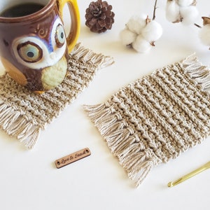 May include: Two beige crocheted coasters with fringe detailing. The coasters are on a white surface with a wooden tag that says "Love & Stitch".