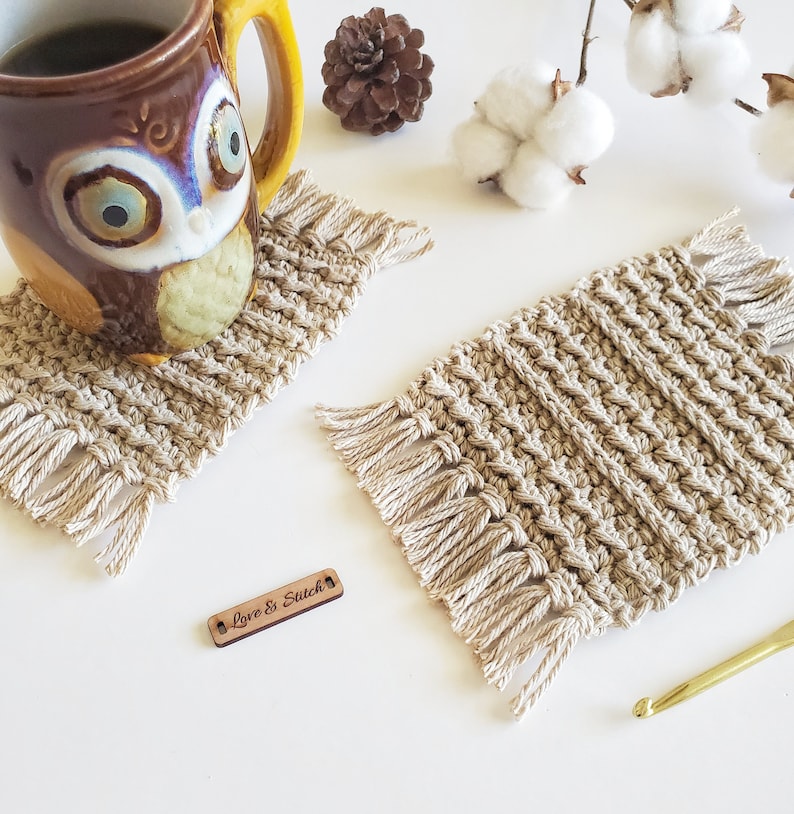 May include: Two beige crocheted coasters with fringe detailing. The coasters are on a white surface with a wooden tag that says "Love & Stitch".