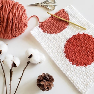 May include: A close-up of a crocheted square with two large red circles. The square is made with white yarn and the circles are made with red yarn. There is a ball of red yarn, a pair of scissors, a crochet hook, a pine cone, and cotton bolls in the background.