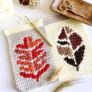 May include: A close-up of a handmade crochet project featuring two leaf designs. One leaf is red and orange, the other is brown and beige. A gold crochet hook, scissors, and yarn are also visible.