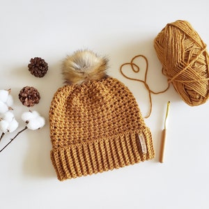 May include: A crocheted beanie hat in a mustard yellow color with a faux fur pom pom on top. The hat has a ribbed brim and a small brown leather label with the text "Crochet Studio" on it. The hat is lying on a white surface with a ball of yarn and a crochet hook.