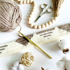 Pode incluir: Um close-up de um projeto de croch&ecirc; com fio marrom e branco, um gancho de croch&ecirc; dourado e uma guirlanda de contas de madeira. Tamb&eacute;m h&aacute; bolas de algod&atilde;o e um par de tesouras na imagem.