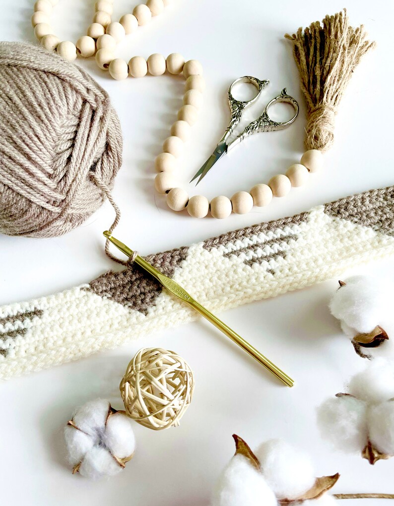 Pode incluir: Um close-up de um projeto de croch&ecirc; com fio marrom e branco, um gancho de croch&ecirc; dourado e uma guirlanda de contas de madeira. Tamb&eacute;m h&aacute; bolas de algod&atilde;o e um par de tesouras na imagem.