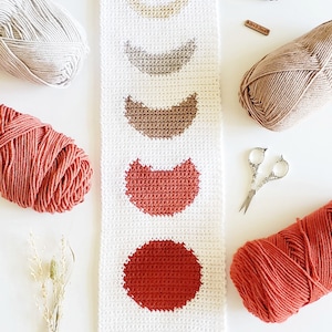 May include: A white crocheted wall hanging with a fringe border features a series of crocheted circles in shades of brown, orange, and red, representing the phases of the moon.