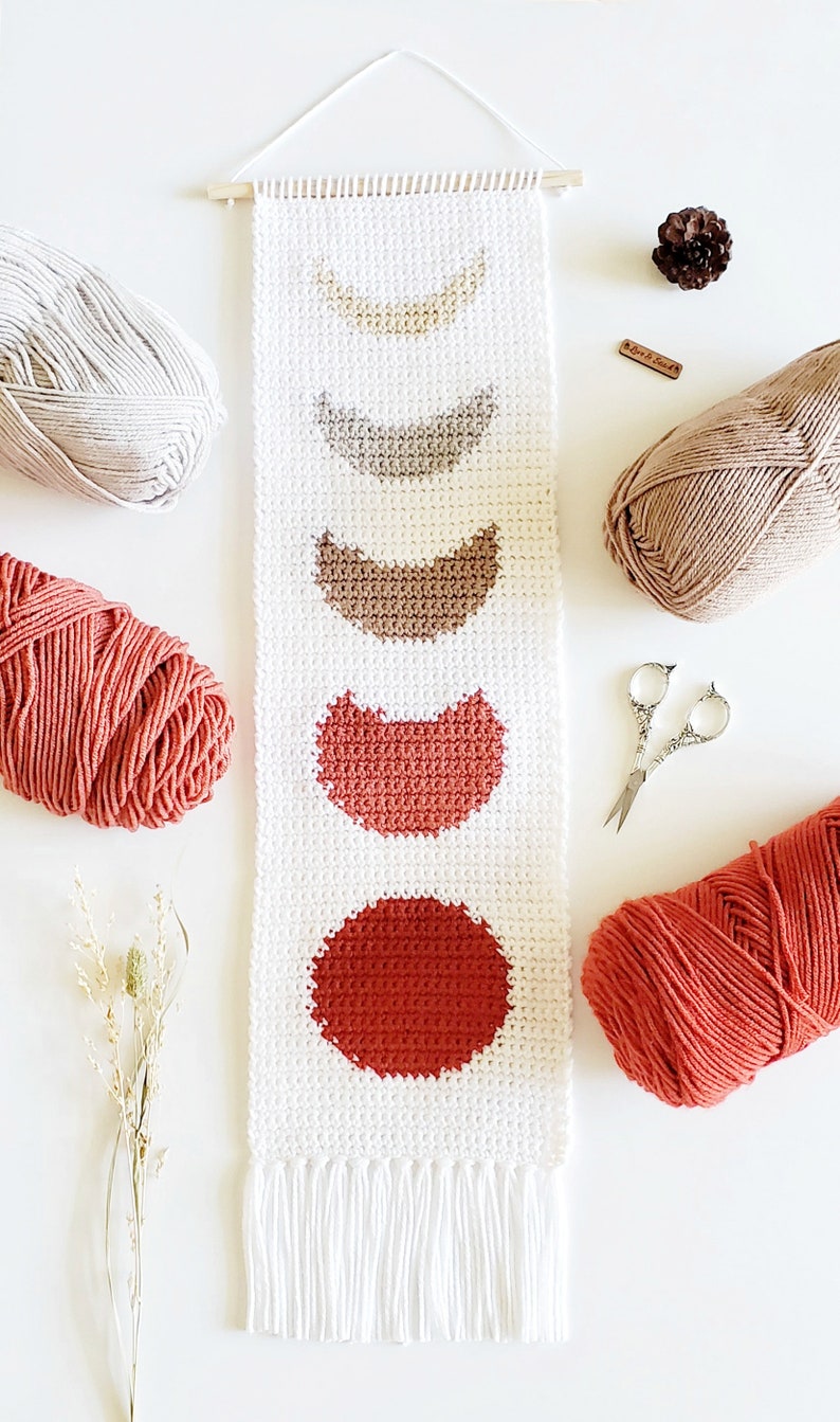 May include: A white crocheted wall hanging with a fringe border features a series of crocheted circles in shades of brown, orange, and red, representing the phases of the moon.