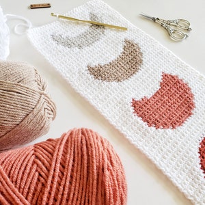 May include: A white crochet project with three crescent moon shapes in shades of beige and coral. The project is partially complete and is lying on a white surface. Two balls of yarn, one beige and one coral, are visible in the foreground.