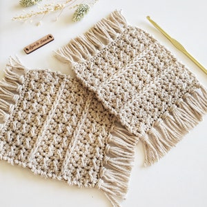 May include: Two beige crocheted coasters with fringe on all sides. The coasters are made with a textured stitch pattern. The text "Love & Stitch" is visible on a small wooden tag.