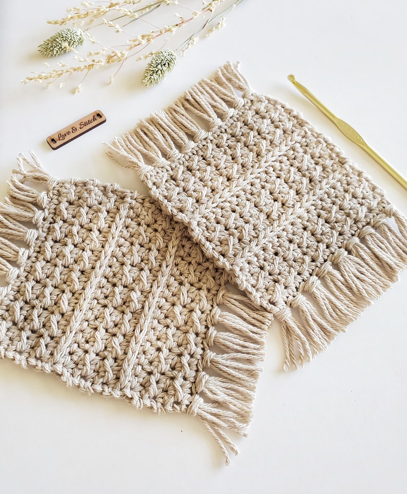 May include: Two beige crocheted coasters with fringe on all sides. The coasters are made with a textured stitch pattern. The text "Love & Stitch" is visible on a small wooden tag.