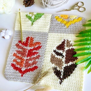 May include: A square, crocheted piece with four leaf designs in shades of red, orange, yellow, green, and brown. The background is a mix of cream and gray. A gold crochet hook, scissors, and yarn are nearby.